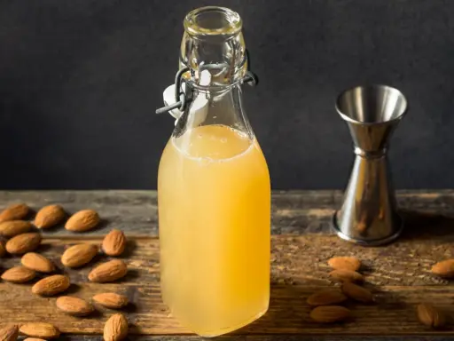Flasche mit selbstgemachtem Mandelsirup, umgeben von Mandeln und einem Messbecher. Perfekt, um Mandelsirup selber zu machen!