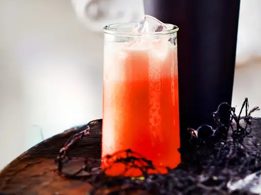 Erfrischender Aperol Sour mit Eiswürfeln in einem Glas, daneben ein dunkler Becher und Spinnweben-Deko.
