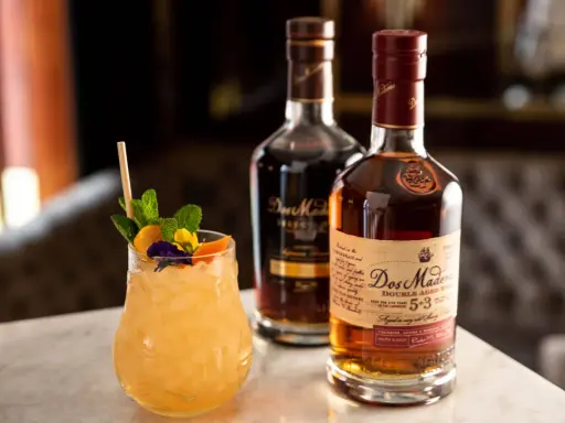 Ein erfrischender Cocktail mit Minze und Früchten neben zwei Flaschen Dos Maderas Rum. Der Alkoholgehalt des Rums ist auf dem Etikett angegeben.