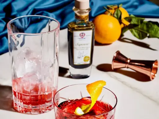 Ein Glas mit Eis und rotem Drink, daneben eine Flasche mit der Aufschrift "Saponoso". Orangen und ein Messbecher vervollständigen die Szene. Grappa, was ist das?