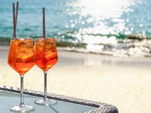 Zwei Aperol Spritz Gläser mit Eis und Orangenscheiben stehen am Strand. Der Aperol alkoholgehalt ist perfekt für einen warmen Tag.
