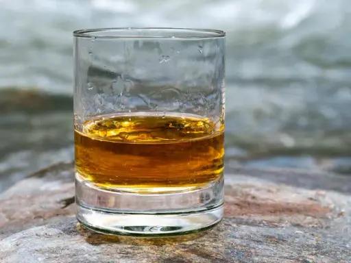 Ein Glas mit bernsteinfarbenem Whisky steht auf einem Stein. Im Hintergrund fließt ein Fluss.