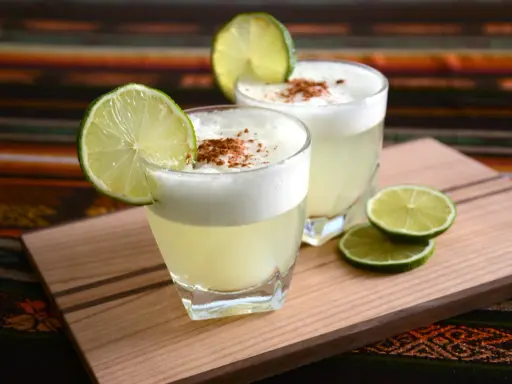 Zwei Pisco Sours, garniert mit Limettenspalten und Zimt, auf einem Holzbrett. Was ist Pisco? Ein peruanischer Traubenschnaps.