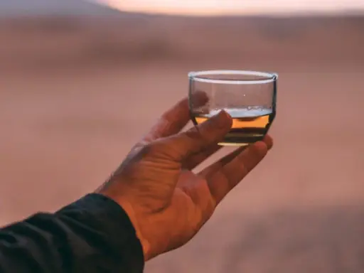 Hand hält ein Glas mit bernsteinfarbenem Rum verschnitt vor einer Wüstenlandschaft bei Sonnenuntergang.
