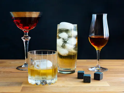 Vier Gläser mit verschiedenen Getränken, darunter ein Glas mit peated whisky, Eiswürfel und Whiskysteine auf Holztisch.