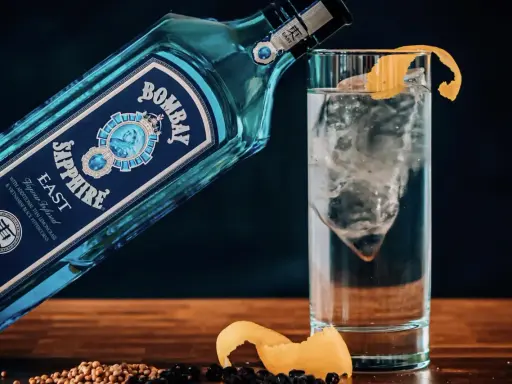 Eine Flasche Bombay Sapphire East Gin lehnt an einem Glas mit Eis und Zitronenschale. Was bedeutet Gin? Erfrischend und aromatisch.