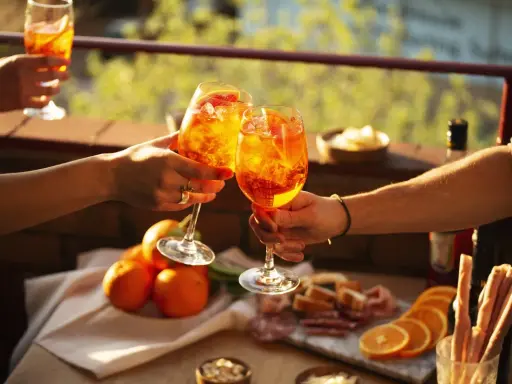 Zwei Hände stoßen mit Aperol-Cocktails an. Der Aperol, aus Rhabarber, Enzian, Chinarinde und Bitterorangen, leuchtet in der Sonne.