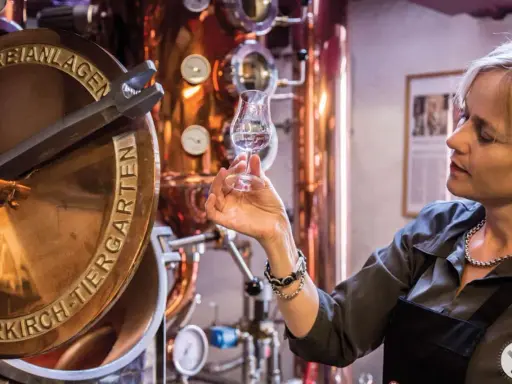 Frau verkostet Genever in einer Brennerei. Kupferne Anlagen und ein Glas mit dem klaren Schnaps sind zu sehen.