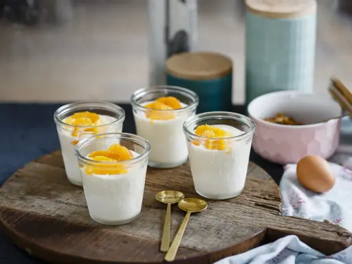 Vier Gläser mit cremigem Dessert, garniert mit Orangenspalten, ideal für Cointreau Rezepte. Zwei goldene Löffel liegen daneben.