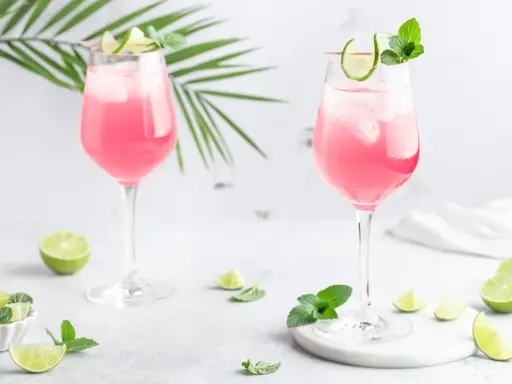 Zwei elegante Gläser mit einem erfrischenden pink spritz, garniert mit Limettenspalten und Minze, umgeben von frischen Limetten und grünen Blättern.