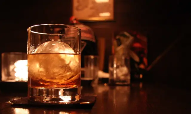 Ein Glas bernsteinfarbener Whisky mit einem großen Eisball. Im Hintergrund sind weitere Gläser und eine gedämpfte Beleuchtung zu sehen, die zum Genuss einlädt, wenn man Whisky mit Eis trinkt.
