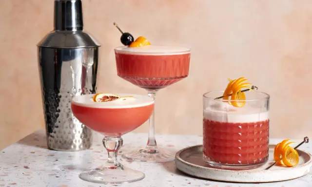 Drei erfrischende Campari Sours, garniert mit Orange und Kirsche, neben einem Cocktail-Shaker.
