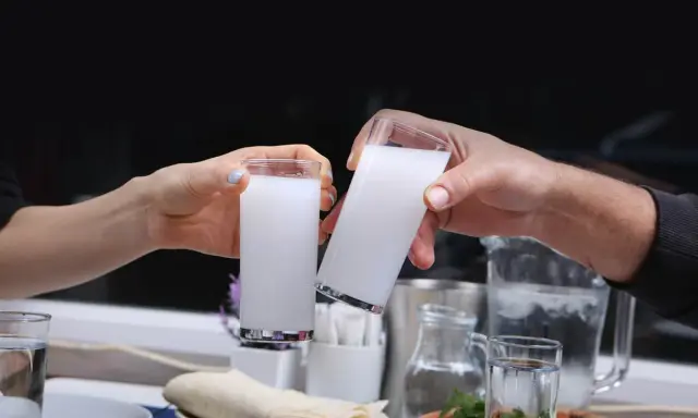 Zwei Hände stoßen mit Gläsern voller weißem Raki an. So trinkt man Raki: langsam, mit Freunden und gutem Essen.