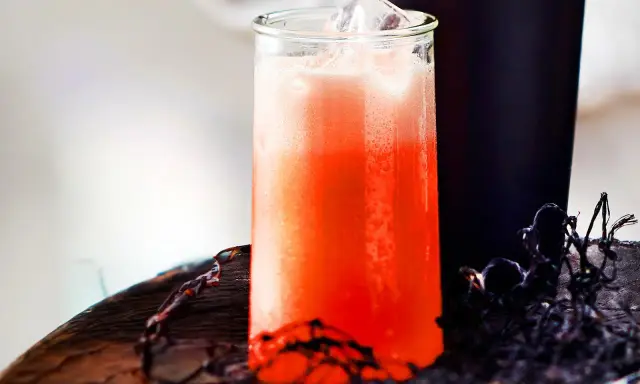 Erfrischender Aperol Sour mit Eiswürfeln in einem Glas, daneben ein dunkler Becher und Spinnweben-Deko.