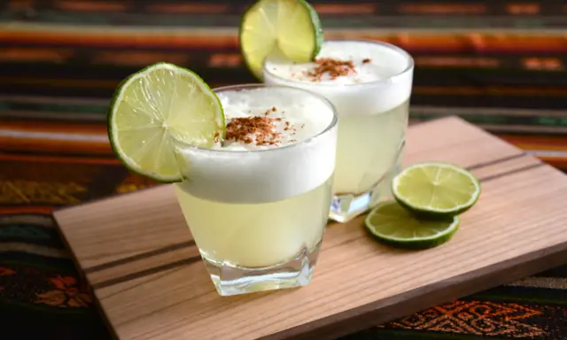 Zwei Pisco Sours, garniert mit Limettenspalten und Zimt, auf einem Holzbrett. Was ist Pisco? Ein peruanischer Traubenschnaps.