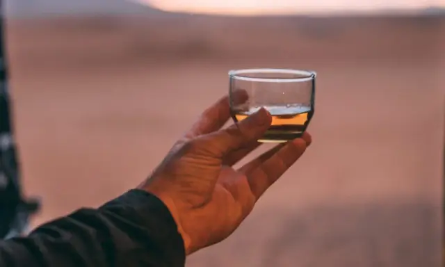 Hand hält ein Glas mit bernsteinfarbenem Rum verschnitt vor einer Wüstenlandschaft bei Sonnenuntergang.