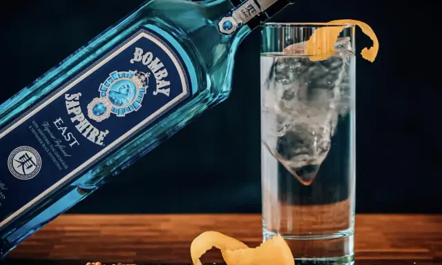 Eine Flasche Bombay Sapphire East Gin lehnt an einem Glas mit Eis und Zitronenschale. Was bedeutet Gin? Erfrischend und aromatisch.