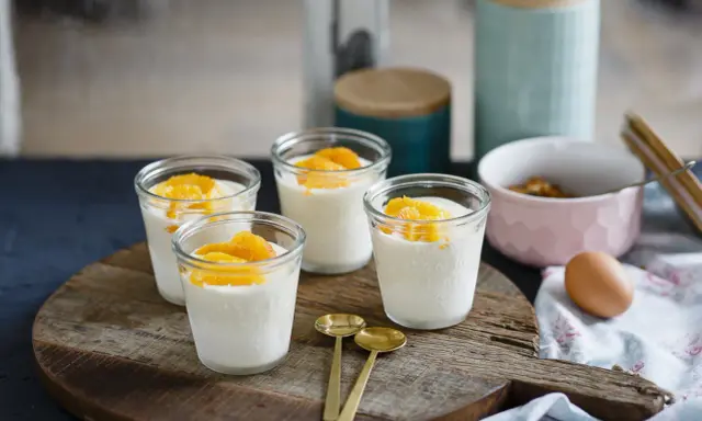 Vier Gläser mit cremigem Dessert, garniert mit Orangenspalten, ideal für Cointreau Rezepte. Zwei goldene Löffel liegen daneben.