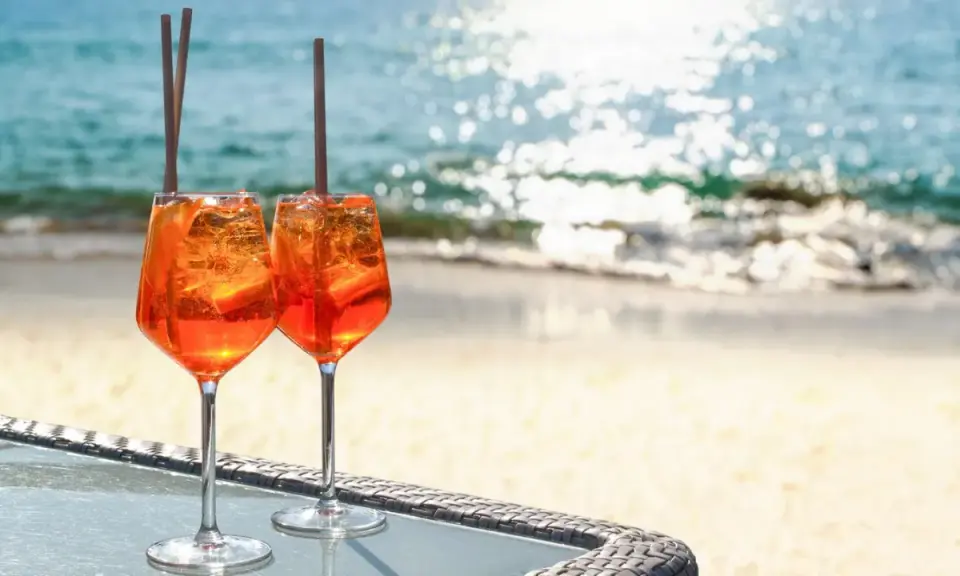Zwei Aperol Spritz Gläser mit Eis und Orangenscheiben stehen am Strand. Der Aperol alkoholgehalt ist perfekt für einen warmen Tag.