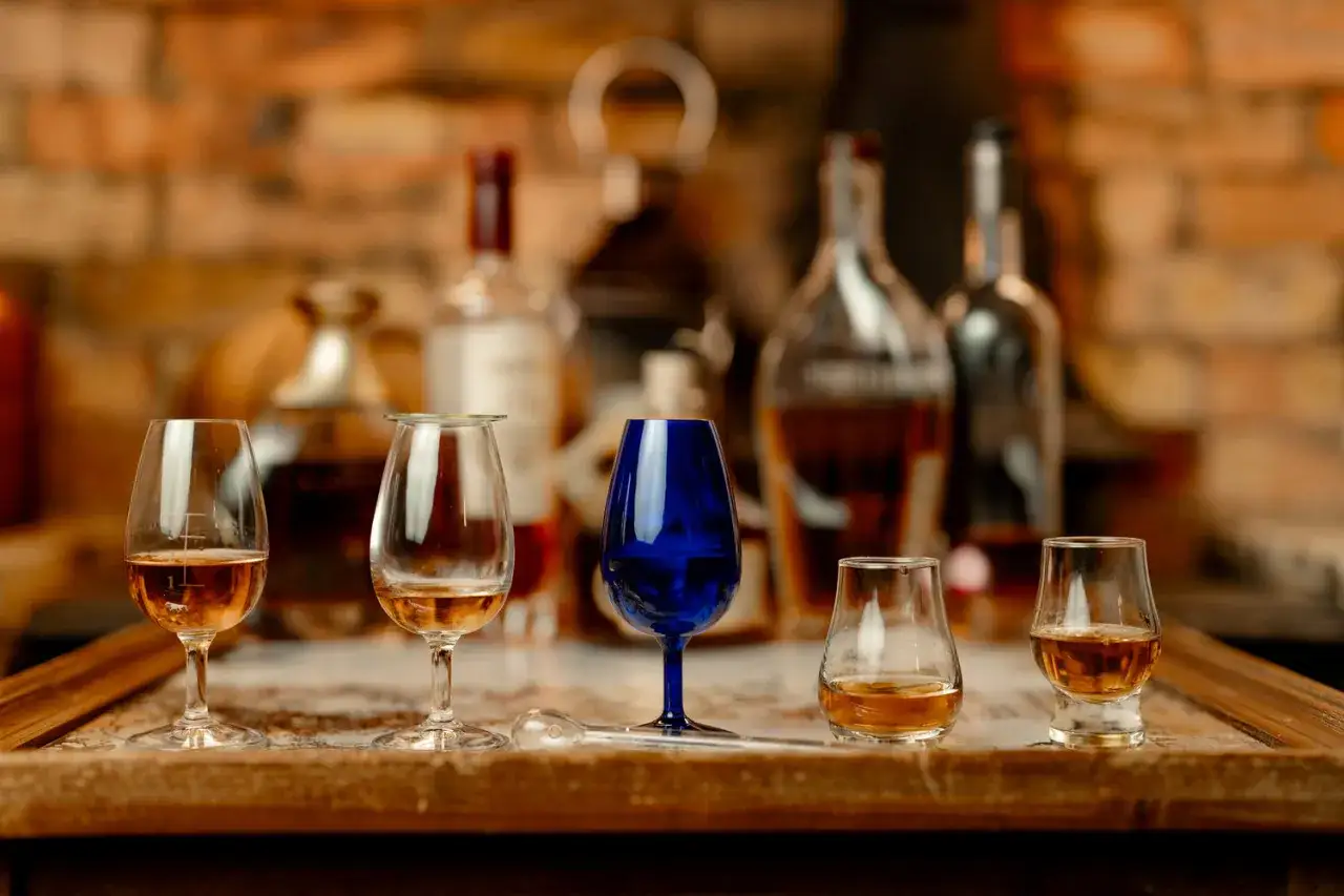 Verkostung von blended malt whisky: Fünf Gläser mit bernsteinfarbener Flüssigkeit stehen auf einem Holztisch vor Flaschen und einer Ziegelwand.