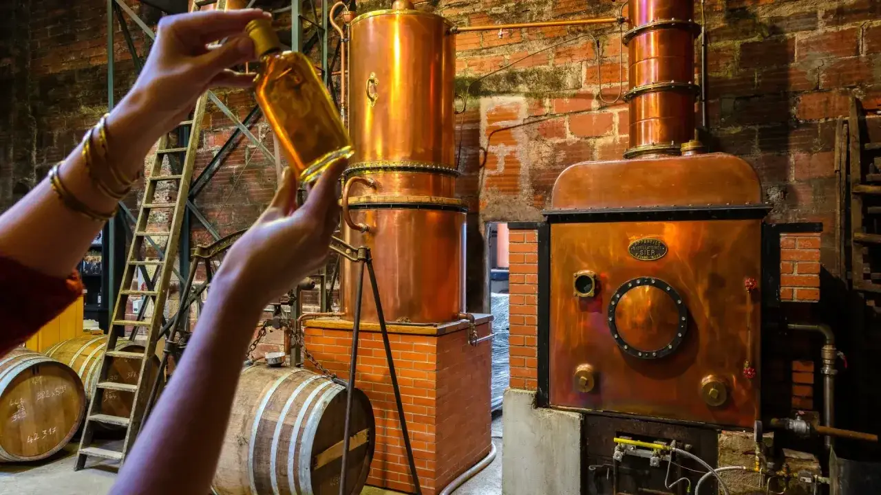 H&auml;nde halten eine Flasche bernsteinfarbenen Armagnac, w&auml;hrend im Hintergrund kupferne Destillierapparate stehen.