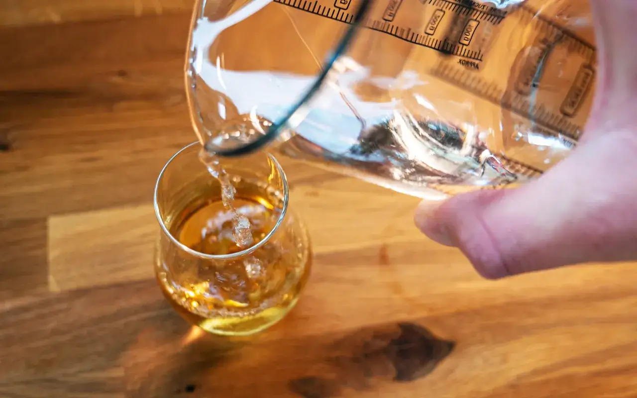 Klares Wasser wird in ein Glas mit bernsteinfarbenem Whisky gegossen. Ein paar Tropfen Wasser ver&auml;ndern den Geschmack des Whiskys.