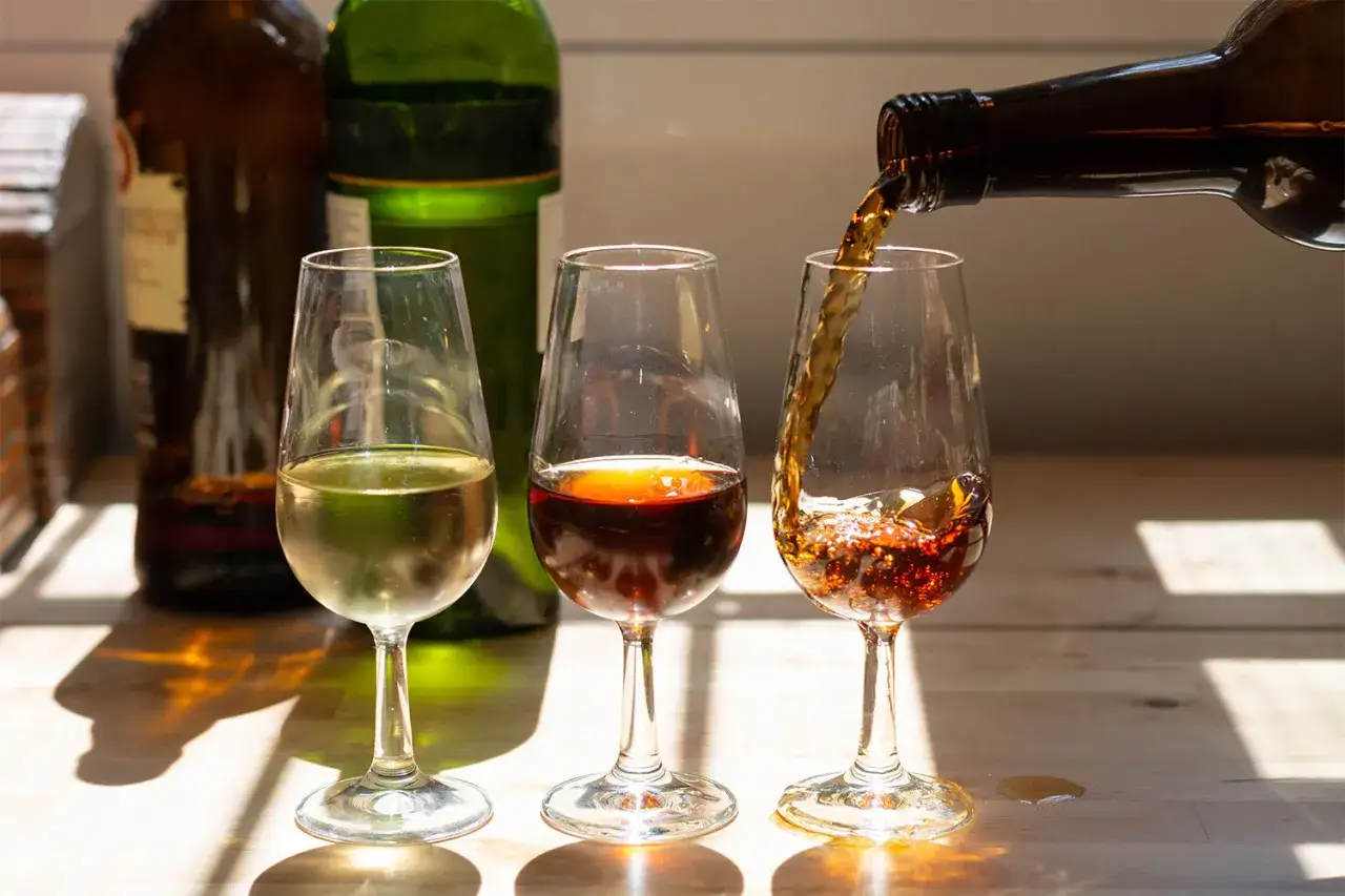 Drei Gläser mit verschiedenen Sherrys: hellgelb, bernsteinfarben und dunkelbraun. Ein Glas wird gerade mit dunklem Sherry gefüllt.