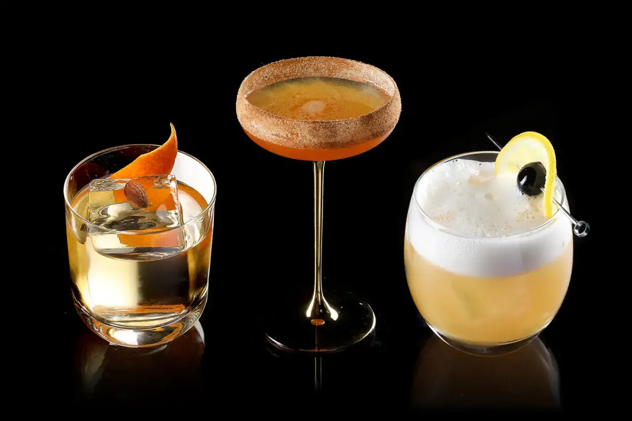 Drei Cocktails auf schwarzem Hintergrund. Links ein Old Fashioned mit Orange und Mandel, in der Mitte ein Amaretto Sour mit Zimtrand, rechts ein Whiskey Sour mit Zitrone und Kirsche.