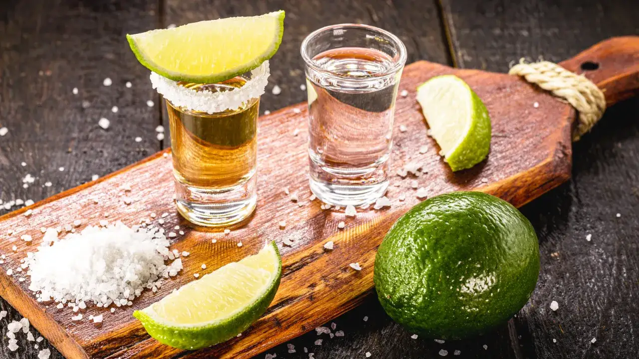 Zwei Shotgläser mit Tequila, eines gold, das andere silber, garniert mit Limettenspalten und Salz auf einem Holzbrett.