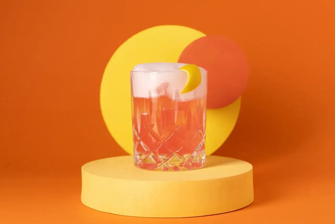 Ein erfrischender Aperol Sour mit schaumiger Haube und Zitronenzeste, präsentiert auf einem gelben Podest vor orangefarbenem Hintergrund mit abstrakten Kreisen.