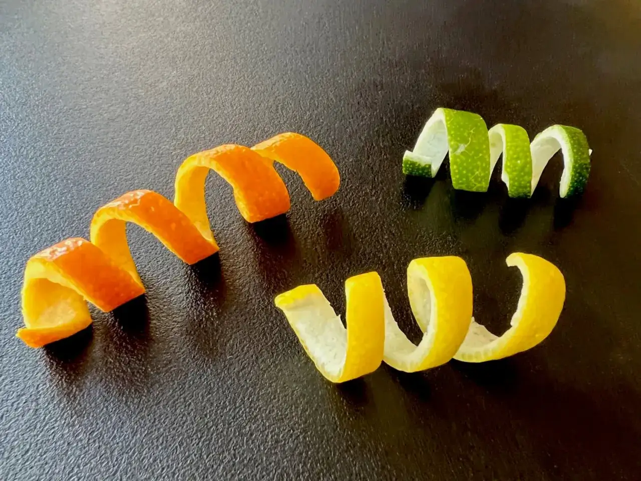 Spiralen aus Zitronen-, Orangen- und Limettenschale liegen auf dunklem Untergrund. Die zitronen zeste duftet herrlich.