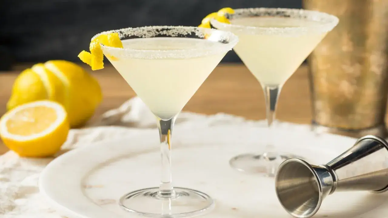 Zwei Martinis mit Zuckerrand und Zitronenzeste, daneben Zitronen und ein Jigger – so serviert man Martini.