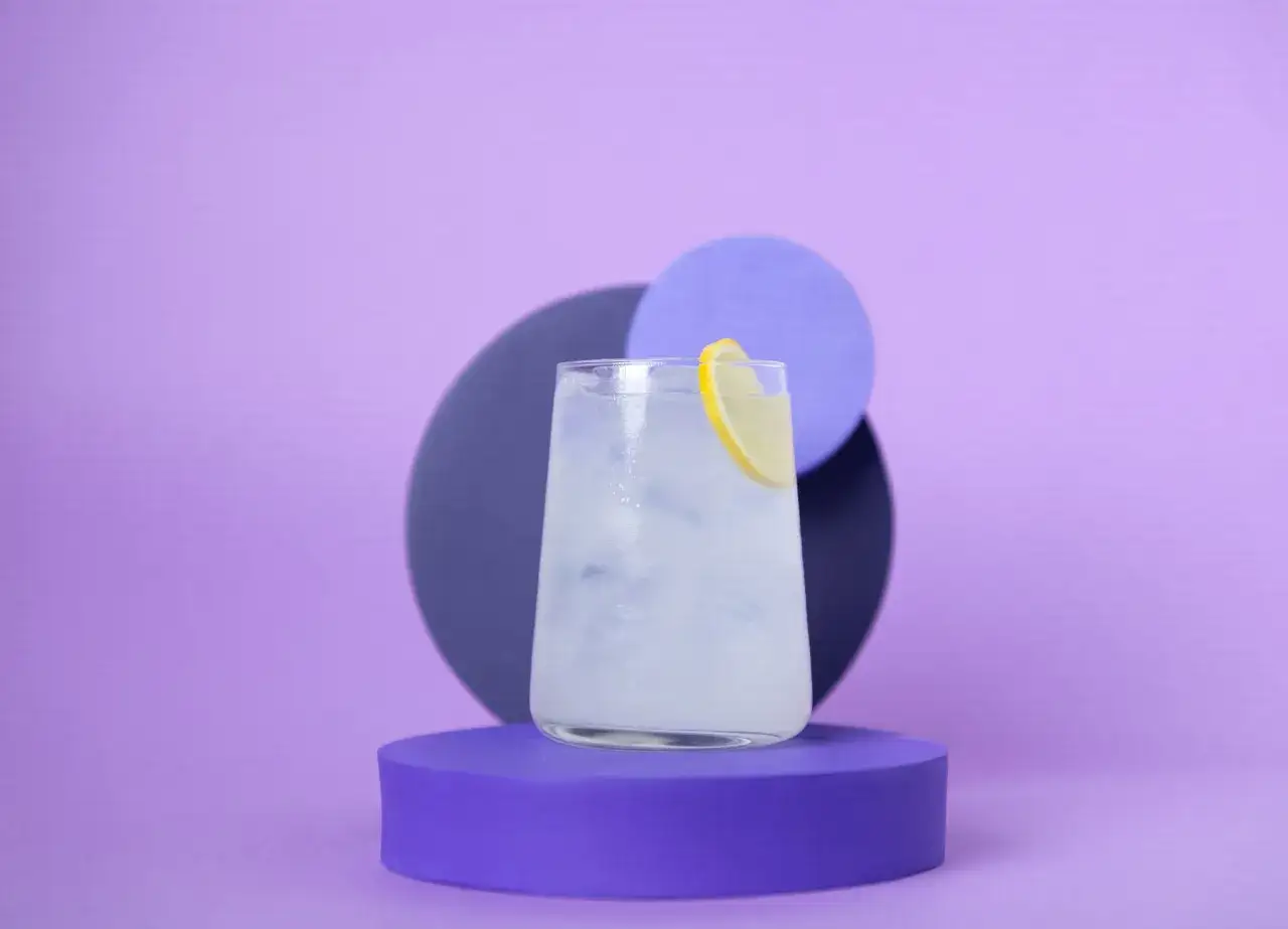 Erfrischender Gin und Bitter Lemon mit Zitronenscheibe, auf lila Podest vor abstrakten Kreisen.