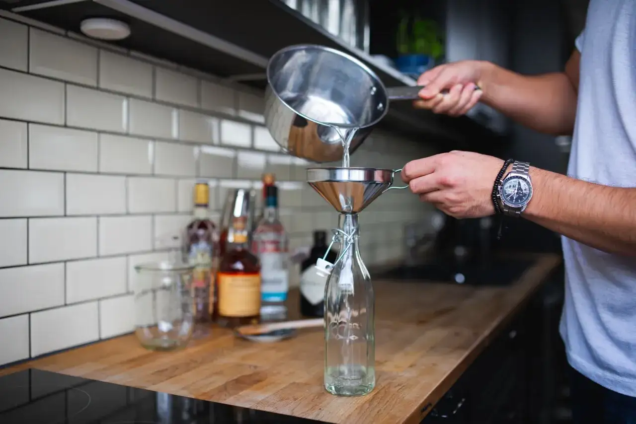 Flüssigkeit aus einem Topf wird durch einen Trichter in eine Flasche gegossen. Das ist ein Schritt bei der Herstellung von Zuckersirup.