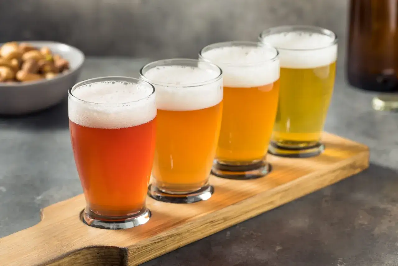 Vier Gl&auml;ser mit Bier, von hellrot bis goldgelb, auf einem Holzbrett. Ein Sch&auml;lchen mit N&uuml;ssen im Hintergrund. Ein Bier mit Gin-Beeren-Geschmack k&ouml;nnte hier gut passen.