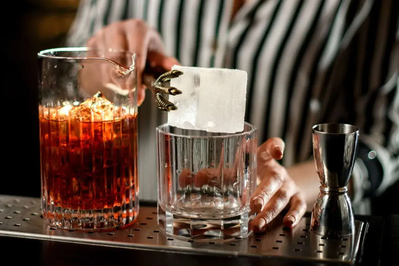Ein Barkeeper bereitet einen Drink on the rocks zu. Ein großer Eiswürfel wird mit einer Greifzange in ein Glas gesetzt. Daneben ein Glas mit bernsteinfarbener Flüssigkeit.