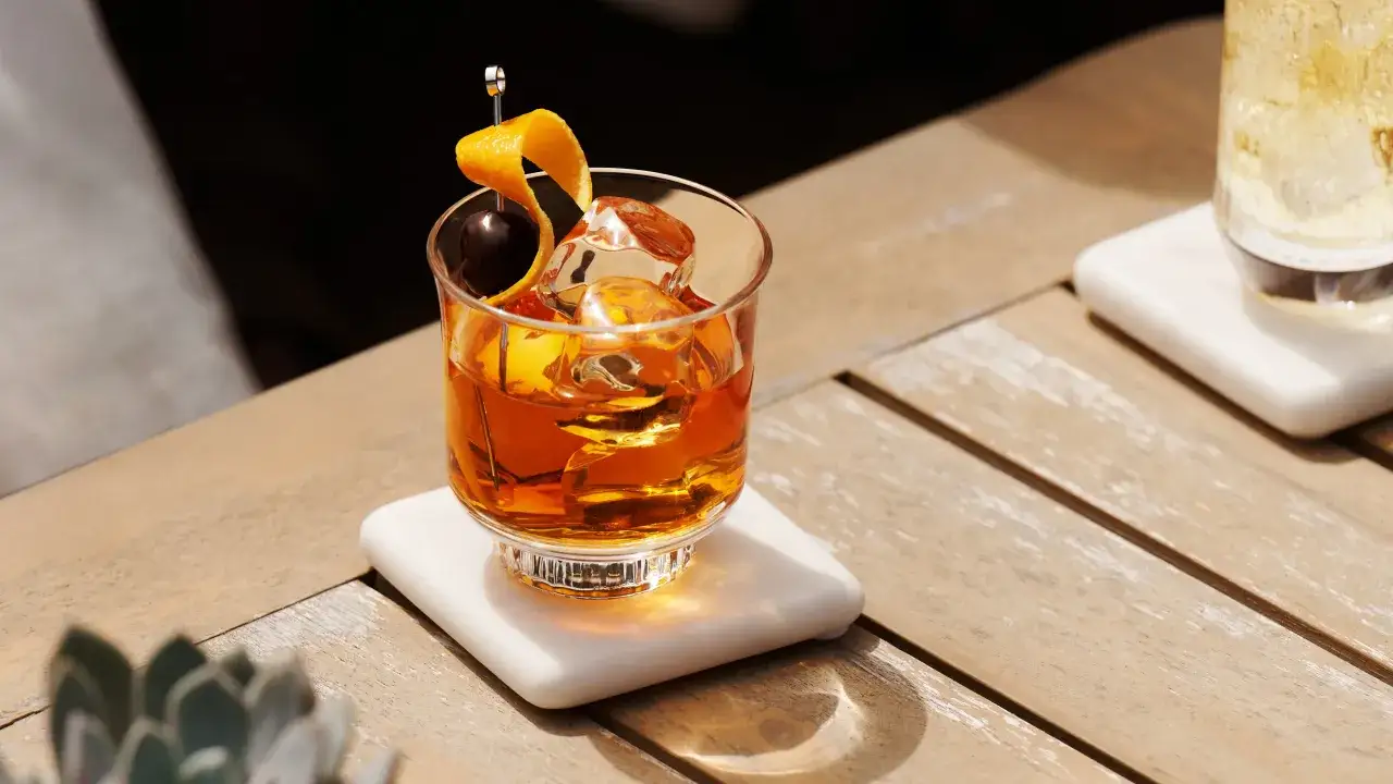 Ein Glas mit bernsteinfarbenem Whiskey neat, Eiswürfeln, einer Kirsche und Orangenzeste auf einem Holztisch.