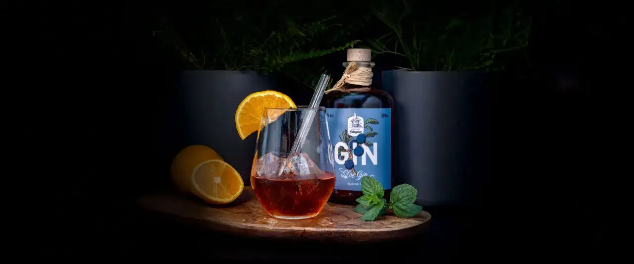 Ein Glas mit Eis, Orangen- und Zitronenscheiben, daneben eine Flasche Sloe Gin und frische Minze.
